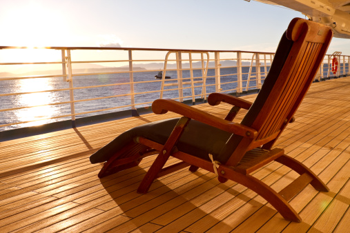 A wooden deck chair is placed upon the wooden deck of a cruise ship.  The chair looks out over the water with sunlight visible on the horizon.