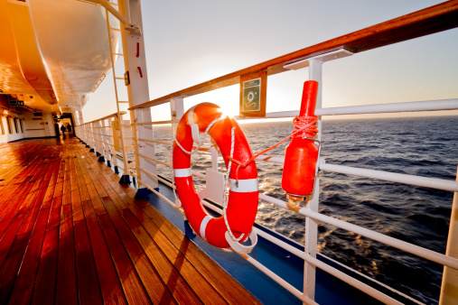 Hanging lifeboat, orange safety beacon and life preserver ring on the railing on the wooden promenade deck of a luxury cruise ship at dusk.  Small aperture creates star effect on the sun as it is peaking from behind the life preserver.  Boat is on a calm blue sea with no land in sight.