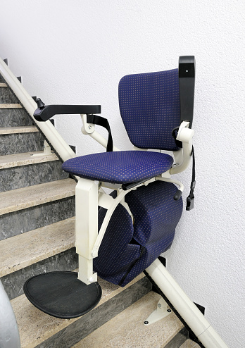 Stair lift mounted to a steep staircase to give disabled or handicapped persons access to a doctor‘s office. The blue chair has a comfortably upholstered seat surface, feet and arm rests and a safety belt for a secure ride. This image shows the stairlift between two levels on its ride upstairs. Vertical orientation.