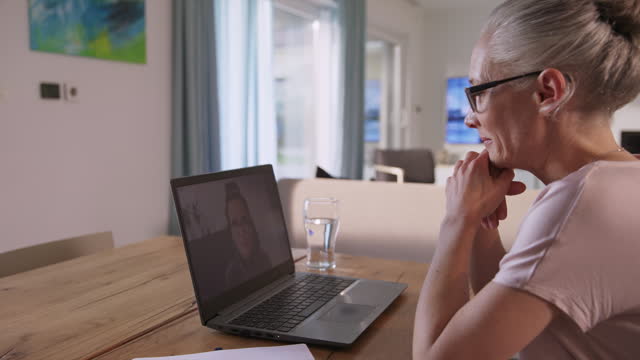 Woman with glasses sitting in her dining room and having a meeting over a videocall on her laptop