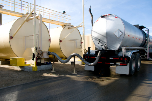 A semi-truck oil tanker is tethered to large oil drums and transfers its oil in to them in this industrial asphalt manufacturing plant.