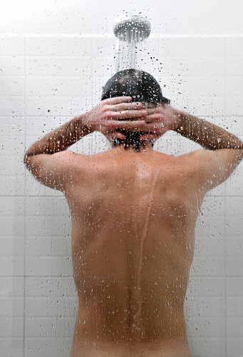 Young, naked man is taking a shower in bathroom, rear view. Shower with white tiles. Simple, modern, interior.