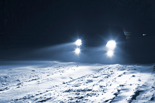 The car goes on a night snow-covered road during a snowstorm. Headlights illuminate the road ahead of the car. View from the side of the external observer. The car on dark-blue background.