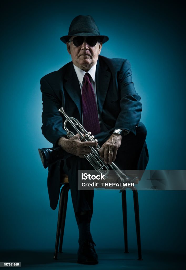 trumpet player resting on a chair mysterious jazz man with a black suit and fedora hat on bluish background posing with his instrument and looking at camera Adult Stock Photo