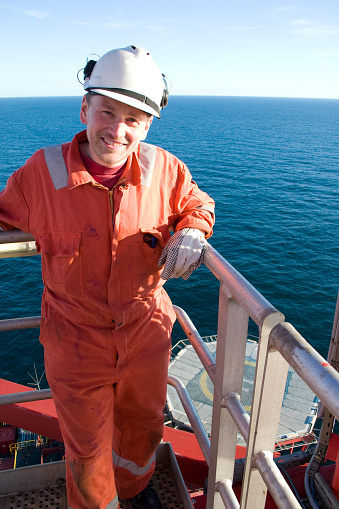 young male roughneck, engineer or technician standing on raised walkway overlooking helideck on an offshore oil rig. Friendly expression good for advertising work, careers, job opportunities and happy work environment. Positive image of work.