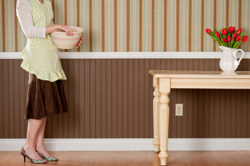 Housewife in apron with mixing bowl. The dining table has red tulips and the wall has a brown beadboard wainscoting and a striped wallpaper.