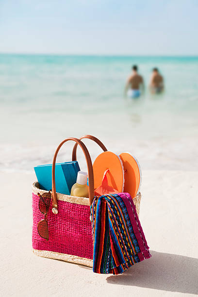 Beach Bag of Summer Gear, Holiday Vacation Suntan Lotion, Accessories A young couple vacationing on their holiday in the Caribbean sea with their beach bag of suntan lotion, towel, book, sunglasses, flip flops and other personal accessories, equipment, and supplies for a tropical beach vacation get-away at Playa del Carmen, near Cancun, Riviera Maya, Mexico. beach bag stock pictures, royalty-free photos & images