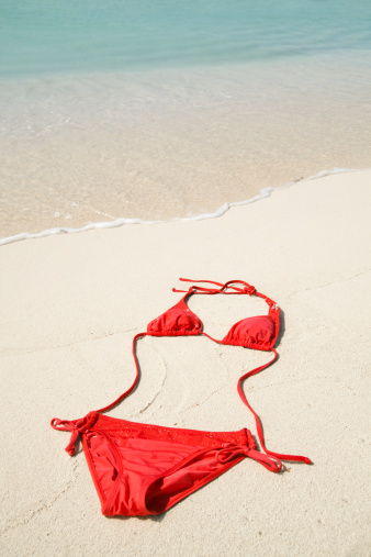 Red bikini swimwear lying next to  water waves on white sandy beach along Caribbean Sea at Cozumel, Mayan Riviera, Mexico. No people are around to claim the empty swimsuit, its strings shaped to form curves of a woman's figure. Playful image for idyllic tropical climate vacation at favorite travel destination.