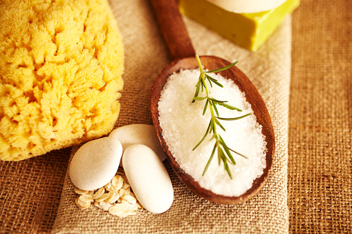 Spa still life of Rosemary on bath salt scrub in wooden spoon on linen, with bath sponge, bars of soap, close-up