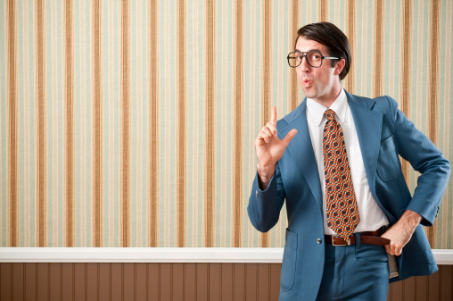 Nerdy businessman wearing a blue retro suit, holding up a smoking gun . The wall has a brown beadboard wainscoting and a striped wallpaper.
