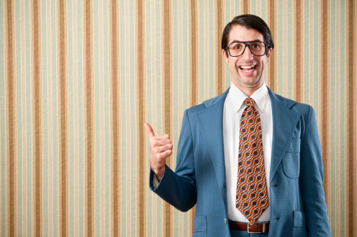 Nerdy businessman wearing a blue retro suit giving a thumbs up. The wall has a brown beadboard wainscoting and a striped wallpaper..