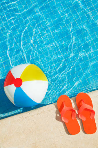 Swimming pool with a beach ball floating in the water and flip flops resting on the pool edge, ready for summer vacation lounging, relaxing, sunbathing, and colorful, cool, wet fun. Vertical still life with dynamic diagonal composition. Outdoor close-up with copy space and no people. 
