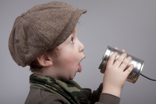 Playful little boy wearing flat newsboy cap holding a tin can phone and making a surprised face, mouth wide open.He is on the left side and faced to right side.The photo was shot in studio with Canon 5D MK2 ISO 100.The background is gray.The hat is used for old fashioned look.