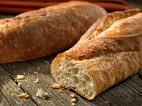 Two fresh baguettes on a wood background. Selective focus is on the cut loaf.