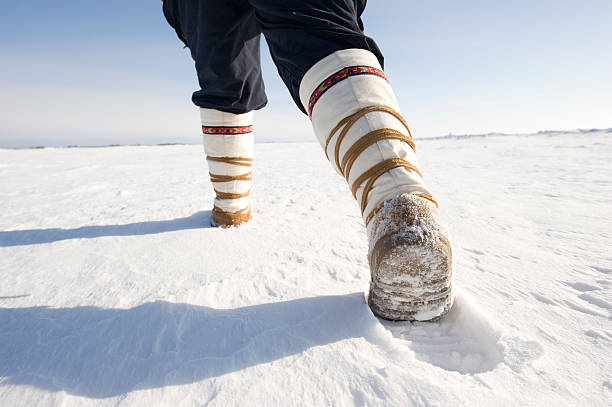 A woman wearing traditional style mukluks (winter boots) strides across the surface of Great Slave Lake in winter.  Low and wide angle shot, heel is raised and good copy space.