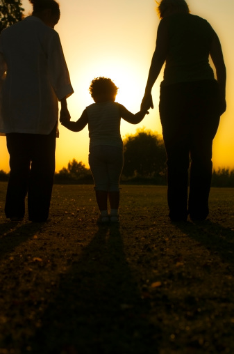 Royalty Free stock photo of child holding hands of her mother and grandmother facing the sunset, shot to create silhouette. Addition of warm effect in post processing and vignette effect.