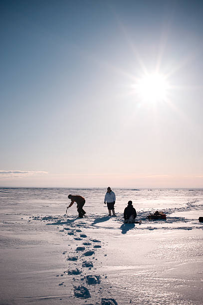 Arctic Ice Fishing, Yellowknife, Northwest Territories, Canada. Three people on the ice of Great Slave Lake drill holes and fish through the ice.  They are backlit.  Lake is snow covered.  Sun is in the sky and good copy space. great slave lake stock pictures, royalty-free photos & images
