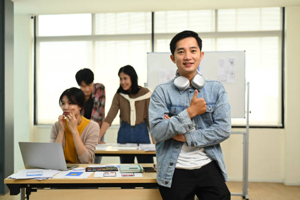 istock Portrait of asian man web developer standing in creative office with his colleagues working on background. 1575744975