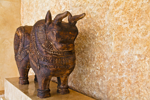 Hand carved bullock from dark indian wood by anonymous artist, probably local indigenous tribal art, hand crafted from probably mango tree, ornate bull freestanding on a plinth against bare mineral tiled wall. This type of craft can be  acquired in local fairs and shops
