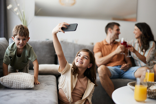 Happy little girl having fun while taking a selfie with mobile phone during time with her family in the living room.