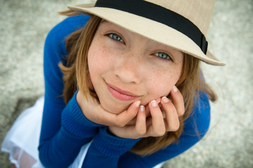 Portrait of a lovely young girl with very blue eyes and a hat sitting down and looking up on the beach. She is wearing a royal blue sweater and white skirt. This was taken in Clearwater, Florida. Shallow DOP, focus on the face. Horizontal shot outdoors in summer.