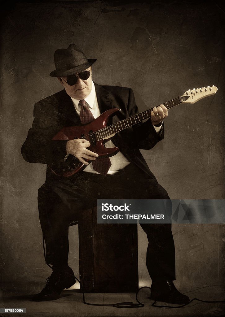 elegant senior playing an electric guitar old school blue- jazz man with a black suit and fedora seating in a wooden crate playing his instrument, composite with grunge papers and vintage textures Adult Stock Photo