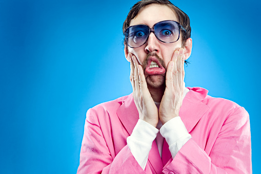A funny looking salesman with a mustache, blue tinted glasses and a pink blazer with a look of horror and shock on his face.  Horizontal on teal blue background.