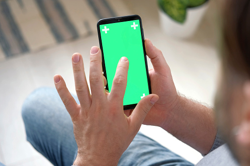 Young man sitting at home holding smartphone green mock-up screen in hand. Male person using chroma key mobile phone. Vertical mode. Touching, swiping display, tapping, surfing Internet social media. Caucasian Man finger clicking, tapping and swiping on center of chromakey green screen. Handsome guy using phone mockup. Close up. Footage Pack. Using for a smartphone, tablet pc or a touch screen devices. Gestures. cellphone with tracking markers. watching content