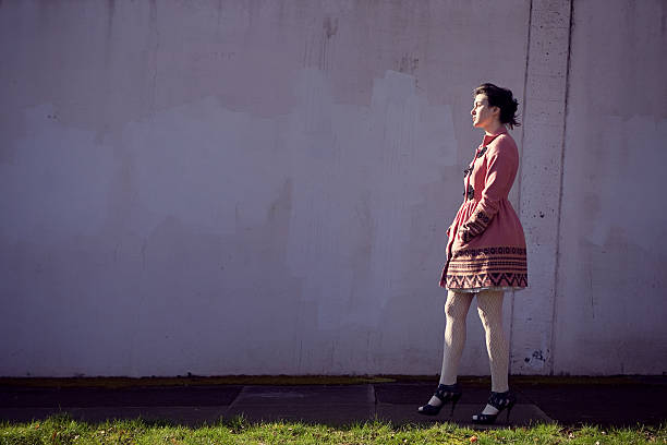 A beautiful young adult woman walks down the sidewalk in front of a large white wall wearing a fashionable retro pink pea coat.  A beam of sunlight  profiles her face; horizontal with copy space.