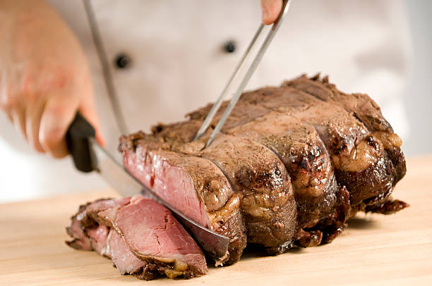 A chef cutting cooked roast beef on a cutting board Professional chef carving slices of a perfectly cooked medium rare prime rib roast beef using a carving knife and fork in a commercial kitchen that could be at a restaurant, hotel, cooking school, café or catering operation in the food and beverage industry. carving set stock pictures, royalty-free photos & images