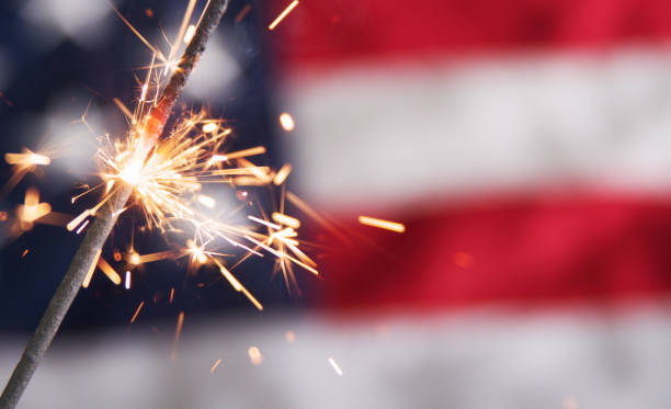 Lit sparkler against a blurred American flag A close-up view of a single burning sparkler. Sparks are flying in front of a partial, blurry view of an American flag in the background. independence day holiday stock pictures, royalty-free photos & images