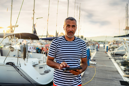 Mature man standing on the dock dressed in a sailor's t-shirt and using a digital tablet