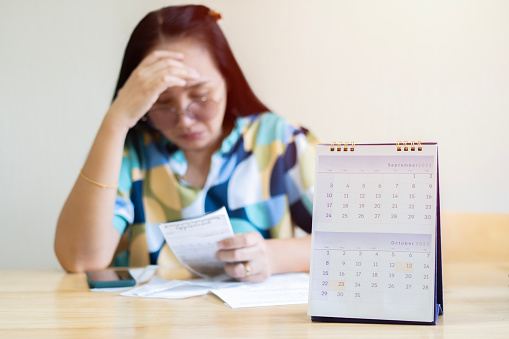 Front view owe Asian woman, female sitting on the chair at home, stressed and confused by calculate expense from invoice or bill, have no money to pay, mortgage or loan. Debt, bankruptcy or bankrupt concept.