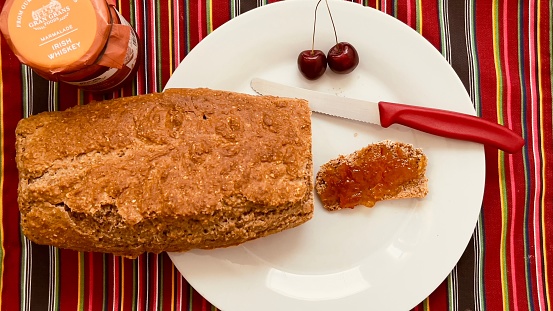 A white plate with a freshly cut slice of bread, jam and a knife on a vibrant tablecloth.