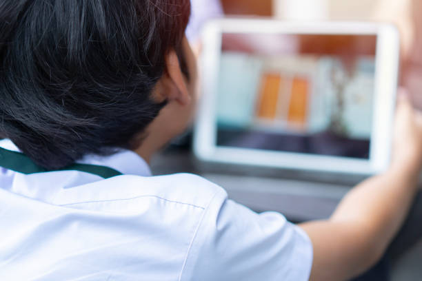 istock Back view of Asian boy using digital tablet while sitting on desk at home. 1575846236
