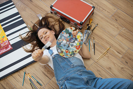 Young dreamy lady holding palette and paintbrush while lying among artist tools in workshop. Talented designer in jeans and white T-shirt creating new project in mind while relaxing on floor indoors.