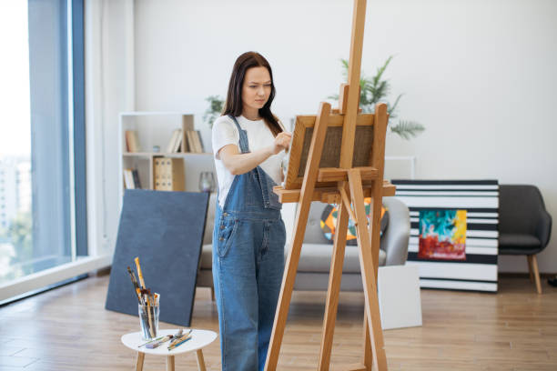 istock Lady with brush and palette posing near canvas on easel 1575853361