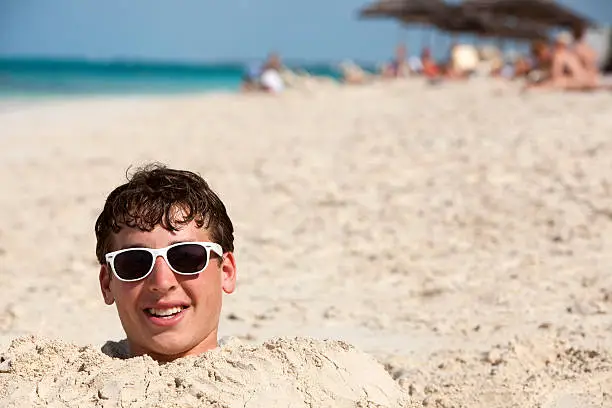 Smiling teenage boy wearing sunglasses is buried in sand on a beach with only his head visible.