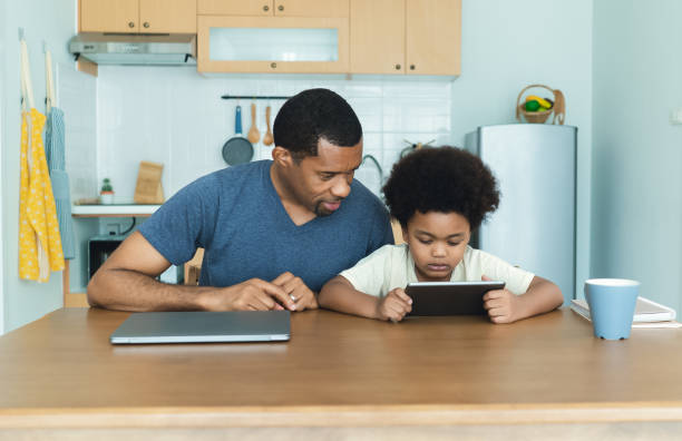 istock Happy African American father and his little son using digital tablet sits together at table in kitchen room. Caring dad and small boy child do home task learn learn online clase. Education concept. 1575863292