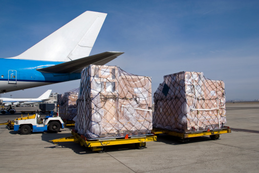 Freight pallets stacked with carton boxes are waiting to be loaded into the aircraft standing behind. The freight transportation airplane can be seen partially.