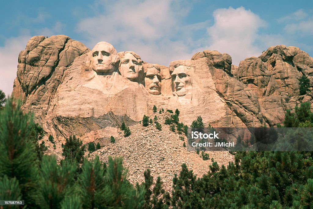 Mount Rushmore on a beautiful summer day. View of Mount Rushmore. The carved faces of the four historical figures, George Washington, Thomas Jefferson, Theodore Roosevelt and Abraham Lincoln are framed by a brilliant blue sky and puffy white clouds. Bright green coniferous trees in the foreground provide contrast to the granite stone into which the faces are carved. Mt Rushmore National Monument Stock Photo