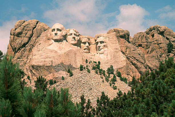 Mount Rushmore on a beautiful summer day. View of Mount Rushmore. The carved faces of the four historical figures, George Washington, Thomas Jefferson, Theodore Roosevelt and Abraham Lincoln are framed by a brilliant blue sky and puffy white clouds. Bright green coniferous trees in the foreground provide contrast to the granite stone into which the faces are carved. custer state park stock pictures, royalty-free photos & images