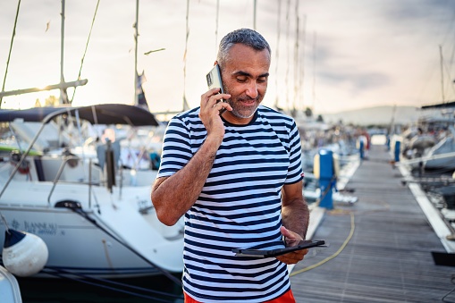 Mature man standing on the dock dressed in a sailor's t-shirt and speaking a phone