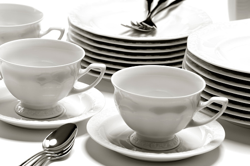 Shot of very elegant china tableware (plates, dessert plates, cups, spoons and forks) on white table. Sepia toned, studio shot.