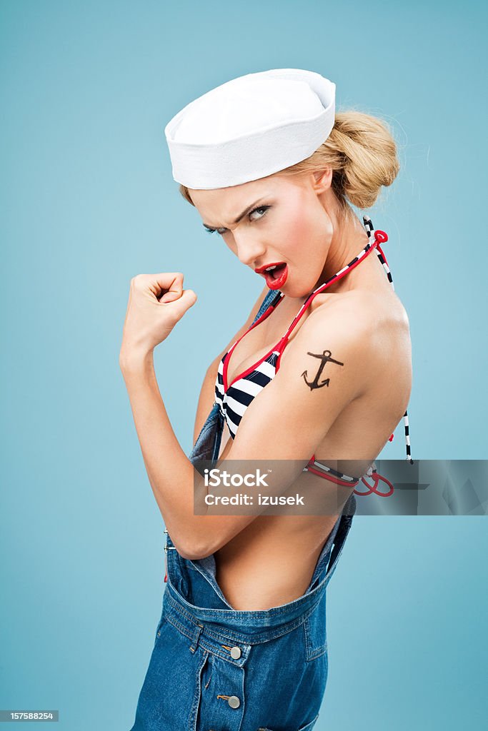Pin-up style sailor woman flexing her arm Young Blond Woman Wearing Striped Bikini and Blue Overalls flexing her arm. Standing against blue background. Pin-Up style. Summer portrait. Muscular Build Stock Photo