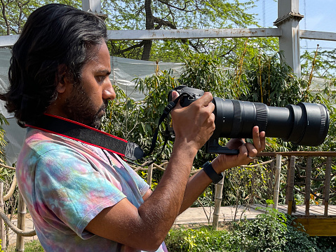 Stock photo showing close-up view of male photographer holding digital SLR camera and using zoom lens to take photographs by looking at camera screen viewfinder.
