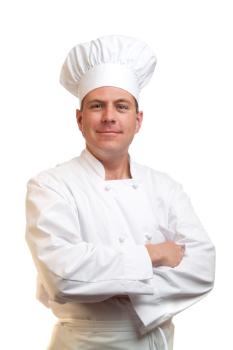 Three-quarter front shot with blurred left side and background of an experienced Caucasian mature baker wearing a white chef's uniform and black apron looking at camera and ready to start his early morning shift at the bakery surrounded by all kitchen supplies.