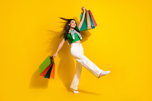 Full length photo of adorable excited lady wear green t-shirt walking rising shoppers isolated yellow color background.