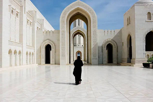 Woman in black Abaya Gown walking towards the giant arch in the famous Sultan Qaboos Grand Mosque in Muscat, Oman, Middle East, Arabia.