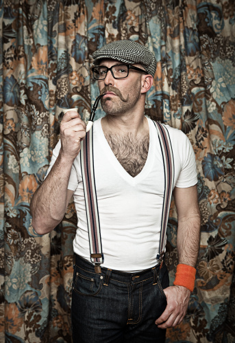 Portrait of an adult male wearing glasses, a poor boy hat and suspenders smoking a pipe while he looks smugly into the camera.